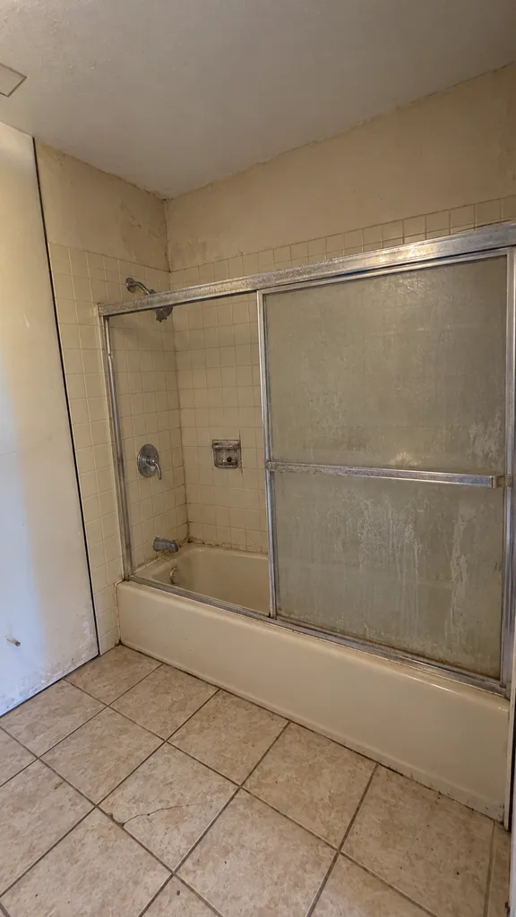 The same bathroom before: an almond fiberglass tub with a cloudy sliding door and dated beige tile